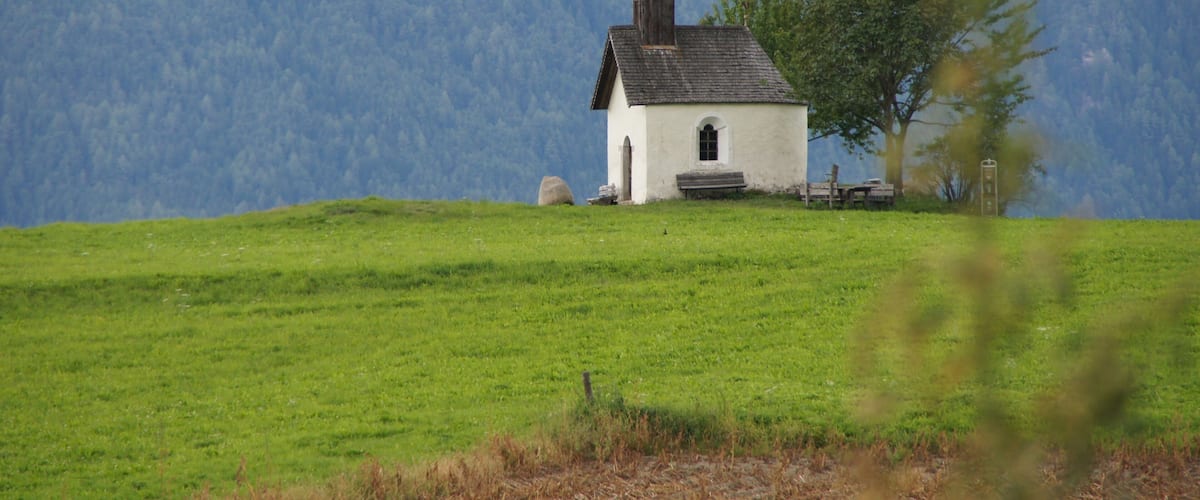 A small white church with a steeple on a grassy hill with a forested mountain in the background.