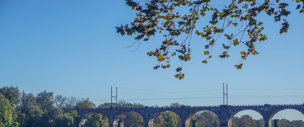 A stone bridge with multiple arches over a calm river, surrounded by trees and clear blue sky.