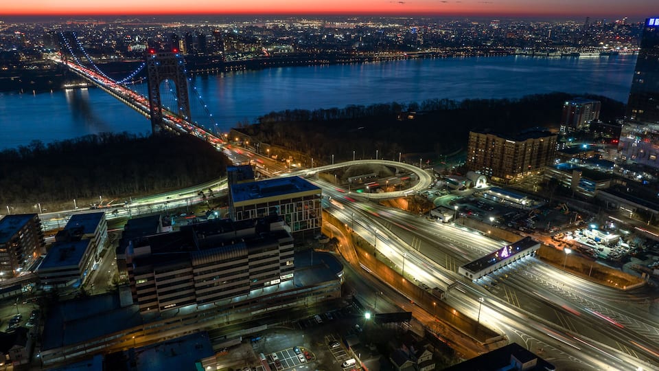 A cityscape at dusk with a prominent bridge, a roundabout, and a river.