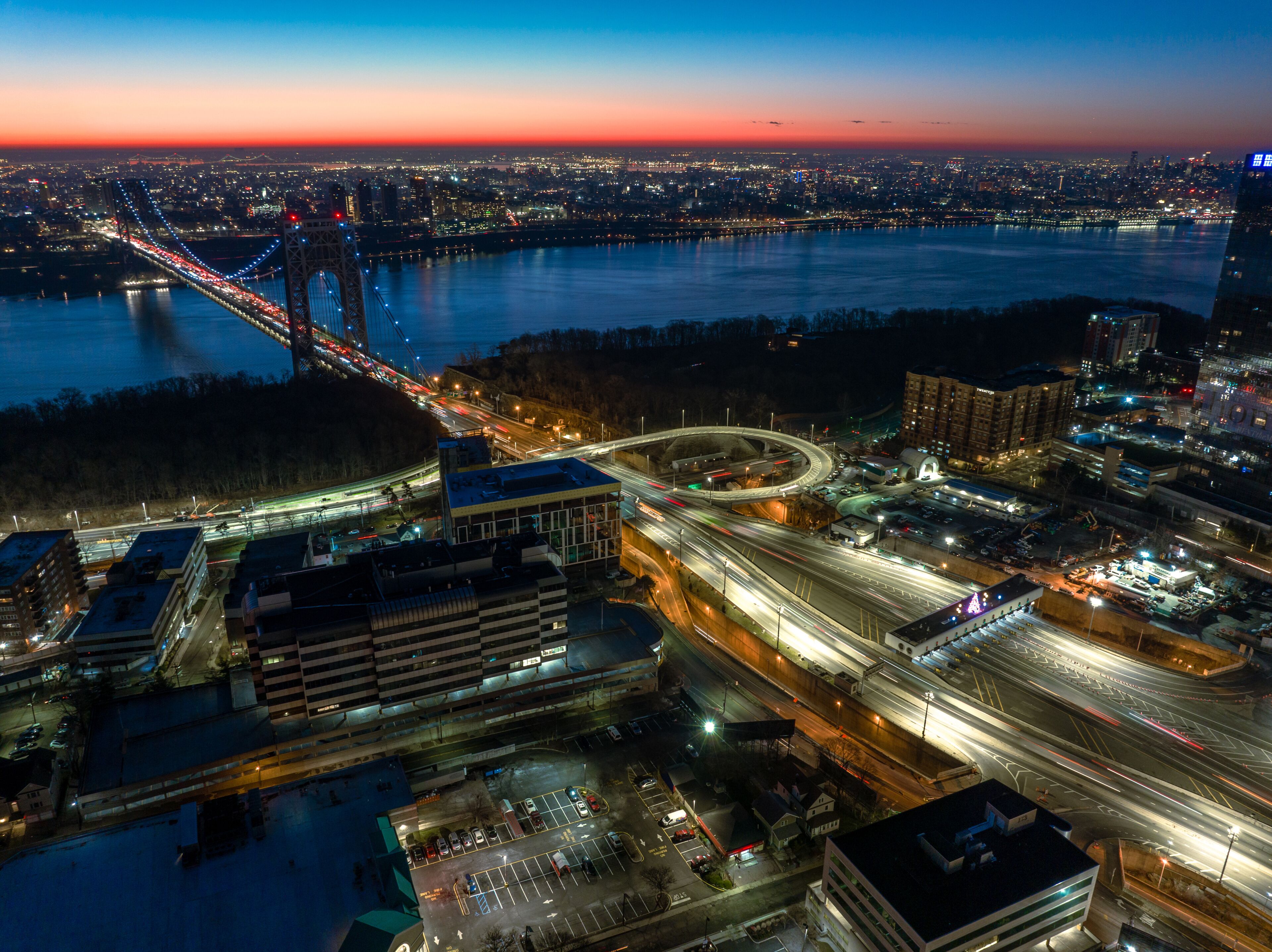 A cityscape at dusk with a prominent bridge, a roundabout, and a river.