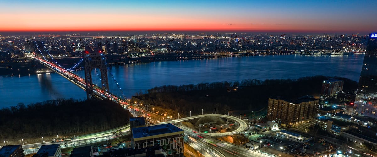 A cityscape at dusk with a prominent bridge, a roundabout, and a river.