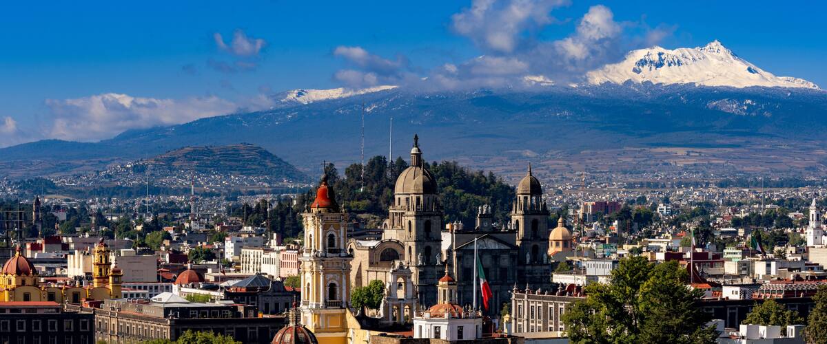 A cityscape with prominent historical architecture, a snow-capped mountain in the background, and a clear blue sky.