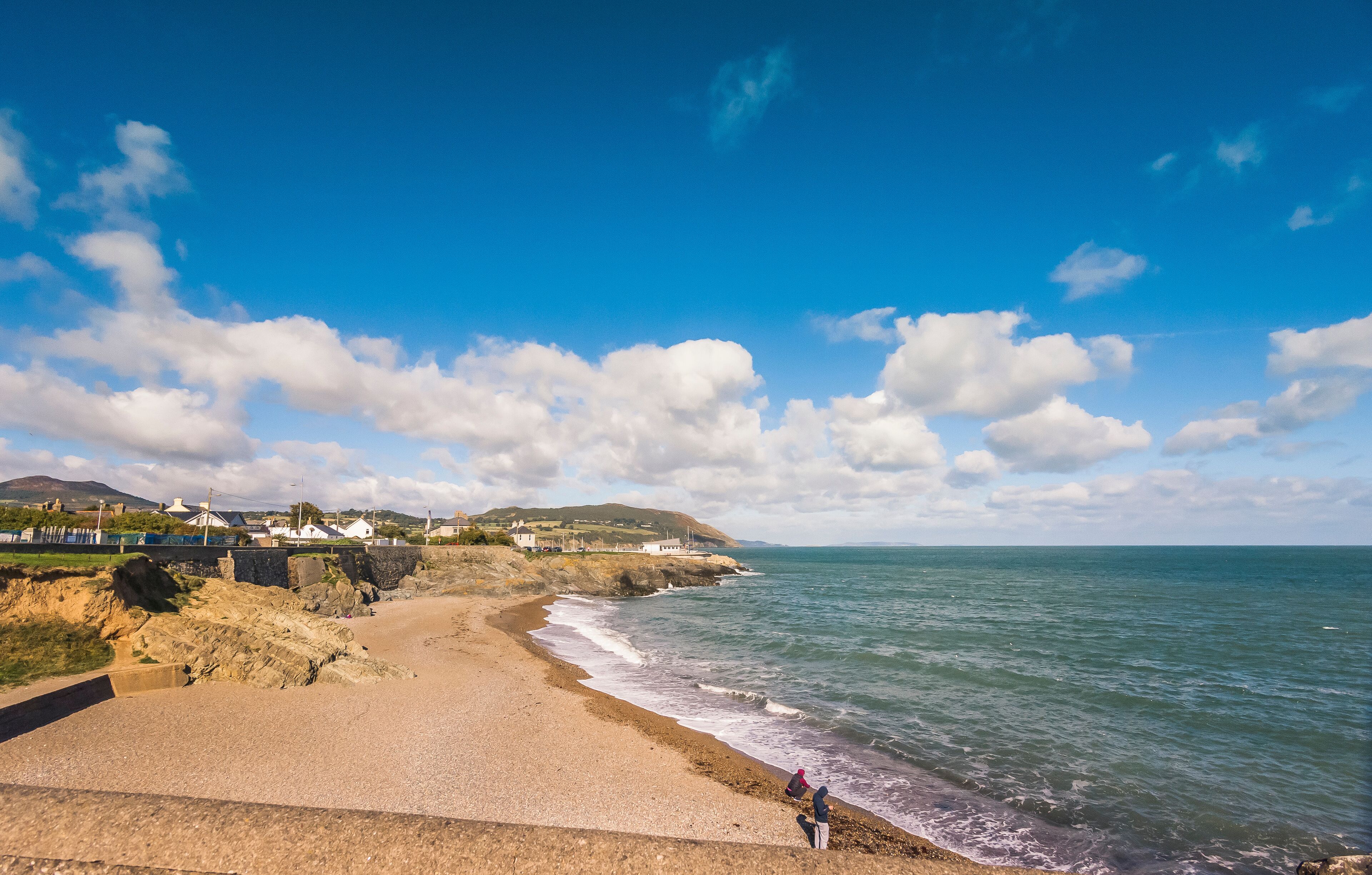 A coastal landscape with a sandy beach, rocky cliffs, and a clear blue sky with scattered clouds.