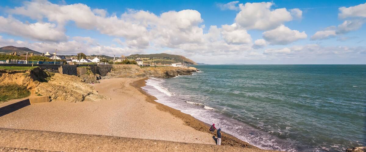 A coastal landscape with a sandy beach, rocky cliffs, and a clear blue sky with scattered clouds.
