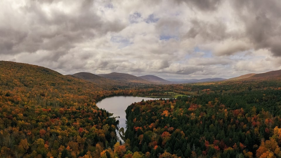 A lake surrounded by autumn-colored forest and mountains under a cloudy sky.