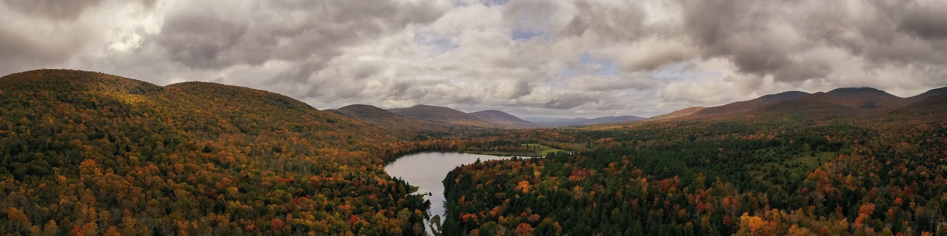 A lake surrounded by autumn-colored forest and mountains under a cloudy sky.