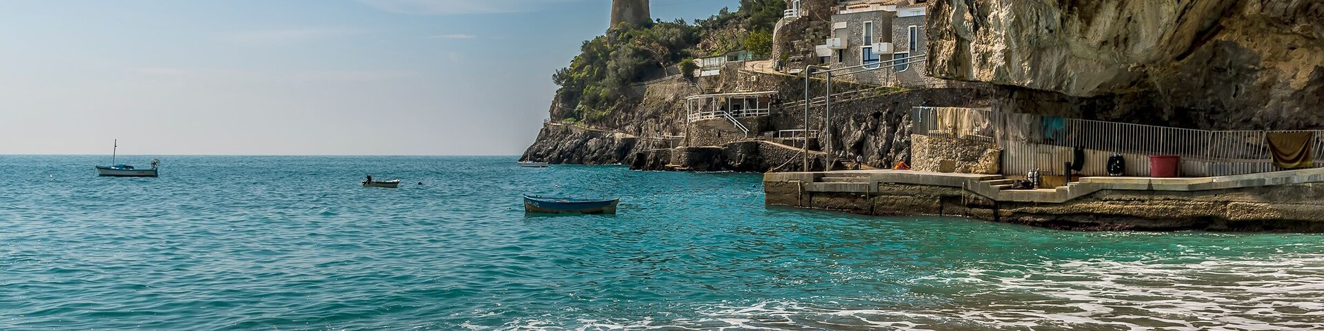 Escena costera con aguas turquesas cristalinas, una playa arenosa y un acantilado con edificios y una torre.