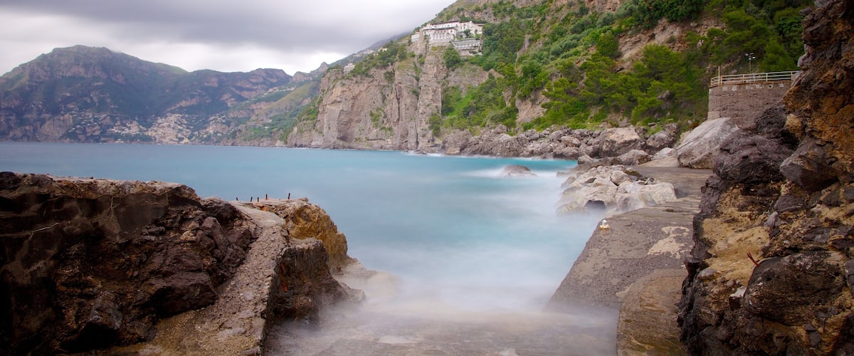 A coastal landscape with rocky cliffs, a clear blue sea, and a building on a hillside.