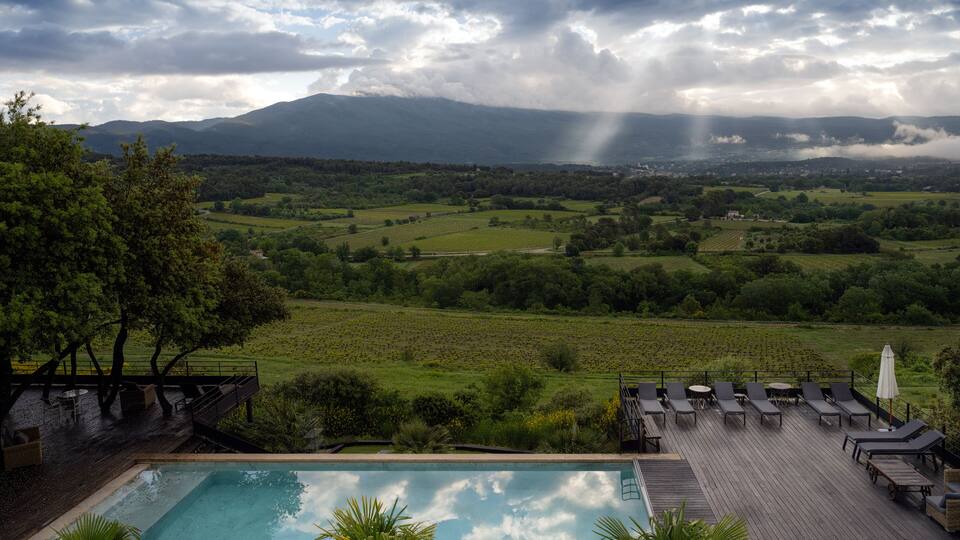 A poolside with lounge chairs, a wooden deck, and a view of a green valley with mountains in the distance.