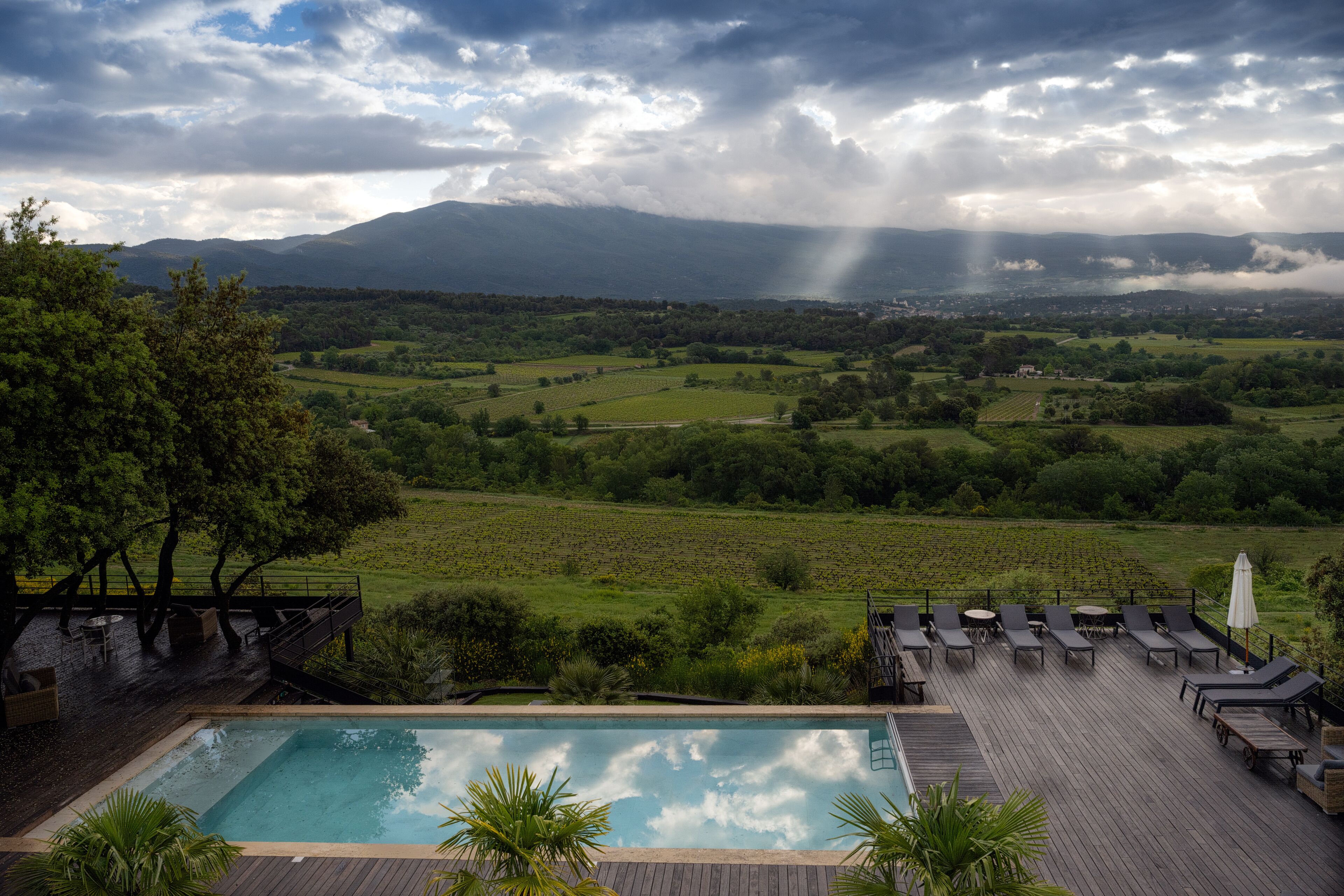 A poolside with lounge chairs, a wooden deck, and a view of a green valley with mountains in the distance.