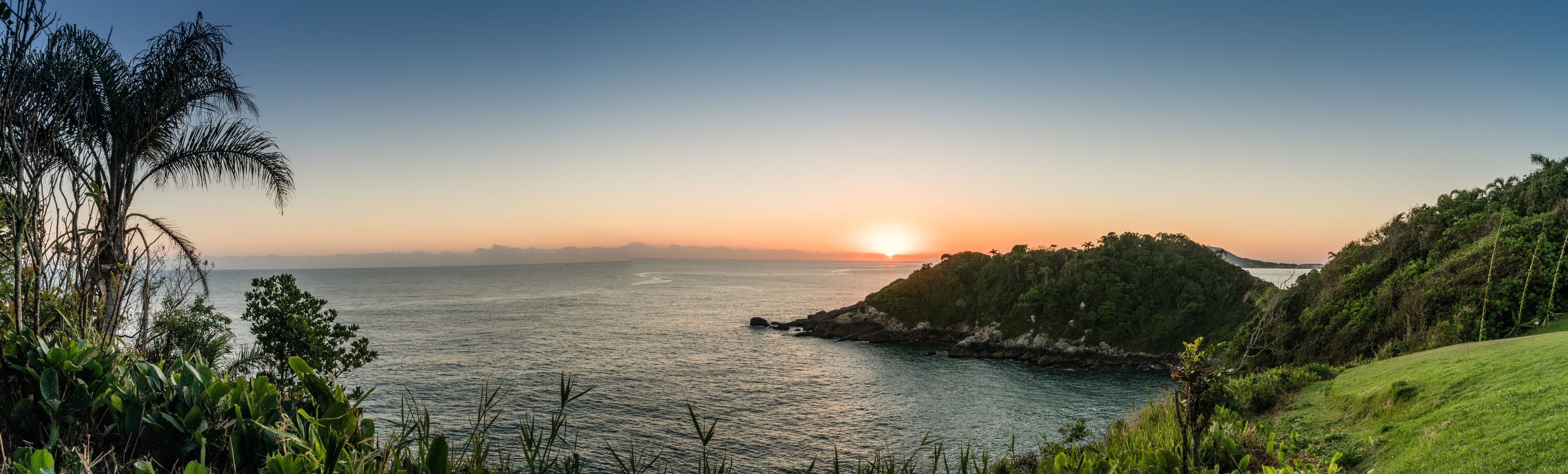 A coastal landscape with a sunset over the ocean, a grassy hill, and dense vegetation.