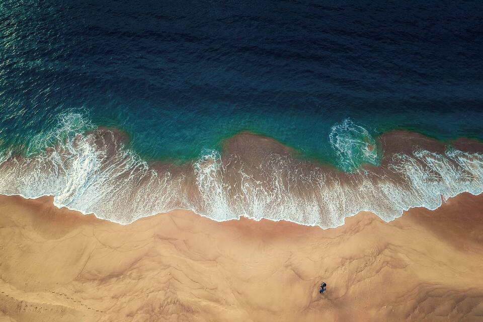 An aerial view of a beach with waves crashing onto the shore and a single person standing in the sand.