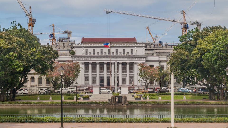 A large government building with columns, a flag, and construction cranes in the background.