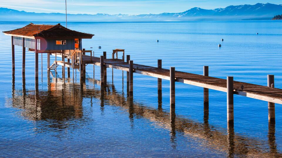 A wooden pier leading to a small house on stilts over water, with mountains in the background.
