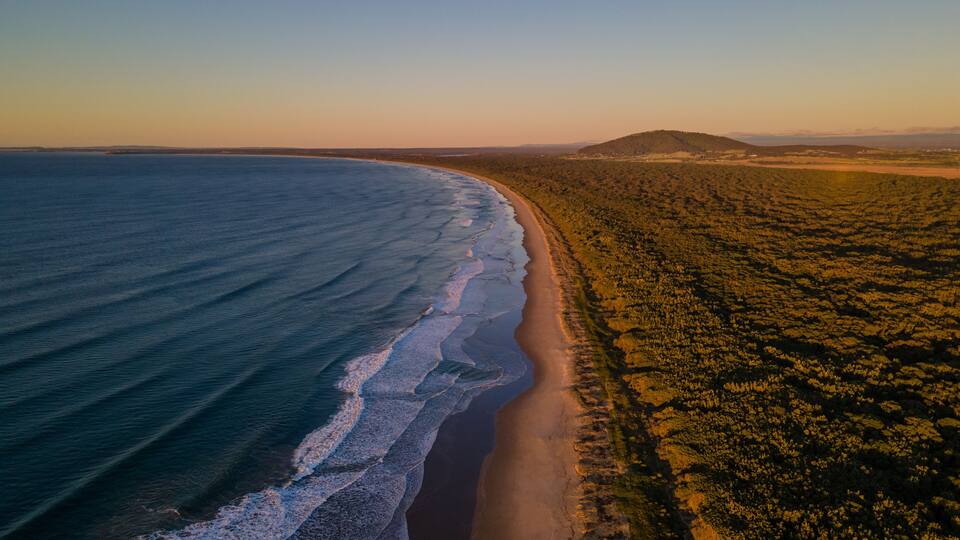 A coastal landscape with a sandy beach, ocean waves, and a hilly terrain in the distance.