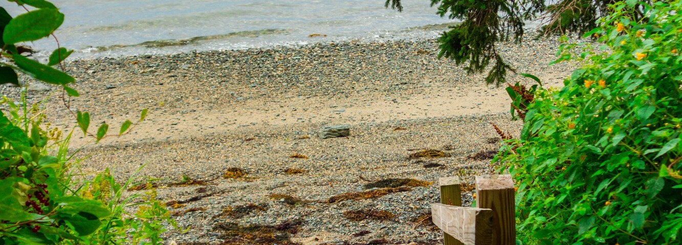 A wooden staircase leading to a pebble beach with a calm body of water and distant land in the background.
