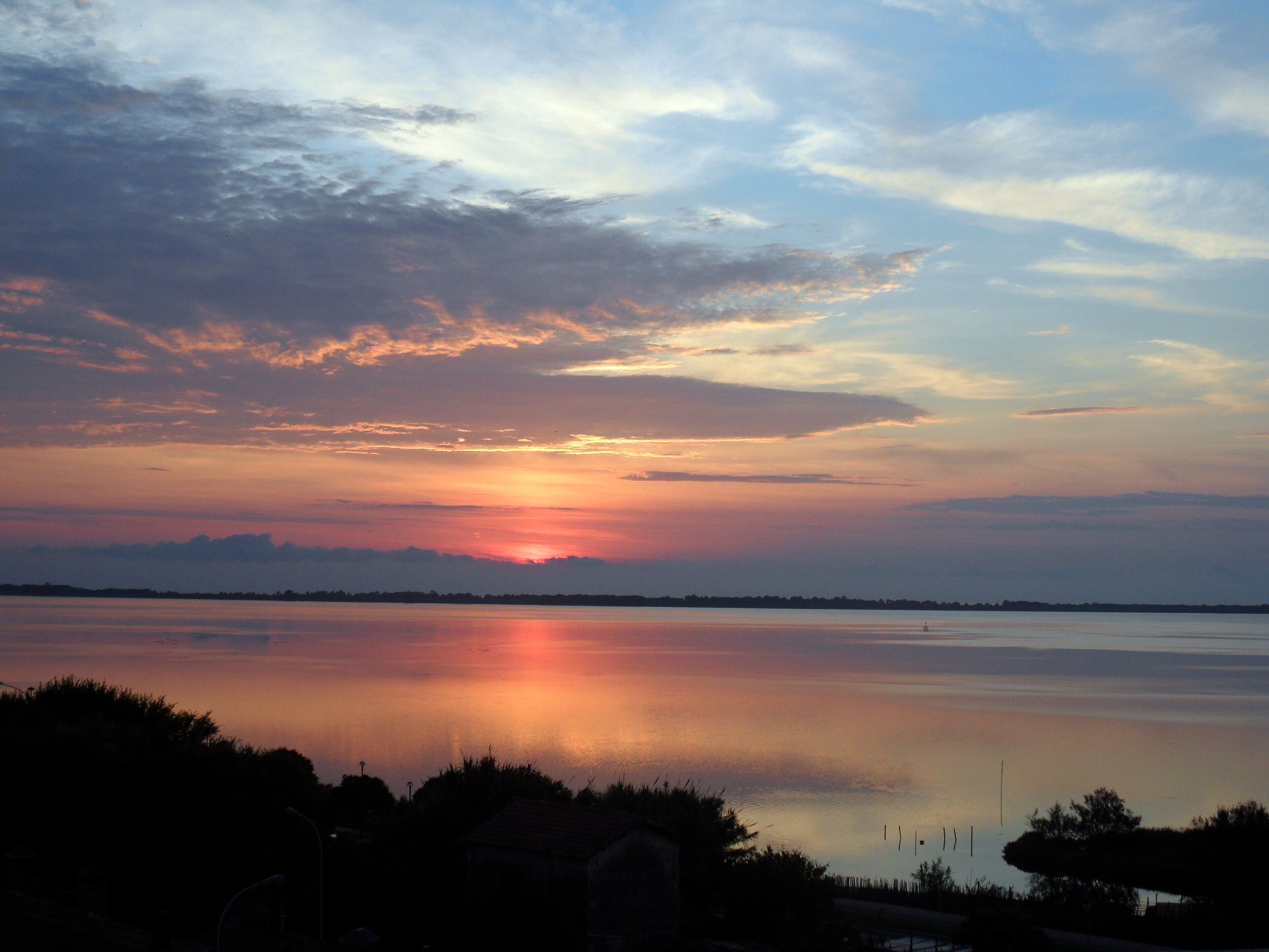 A sunset over a calm lake with silhouettes of trees and a distant shoreline.