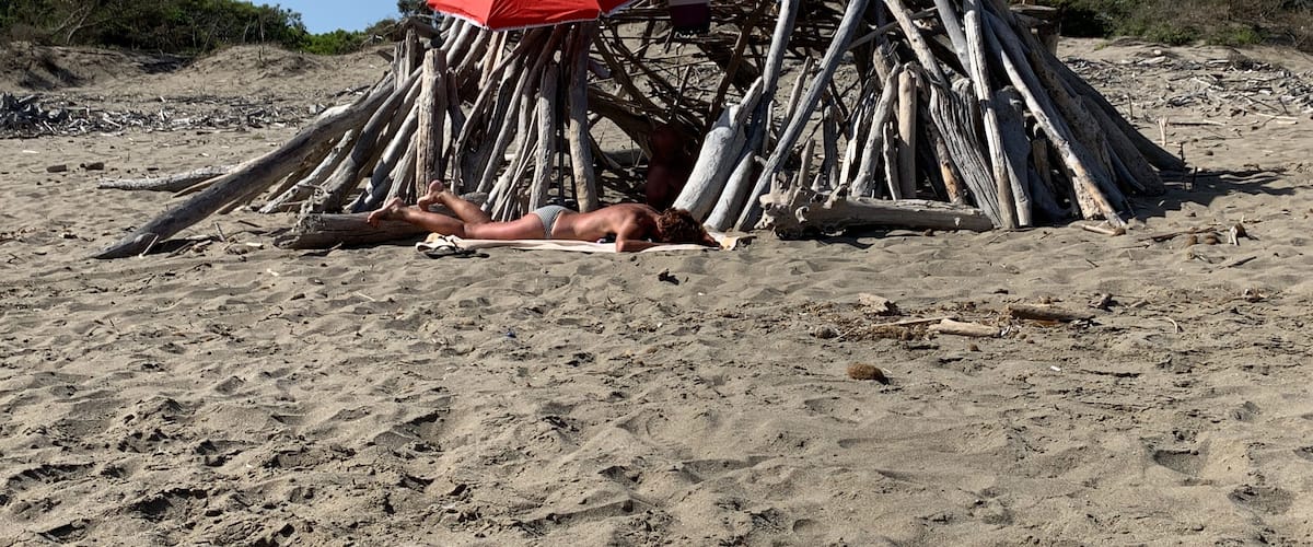 A person lying on a beach under a makeshift shelter made of driftwood and branches.