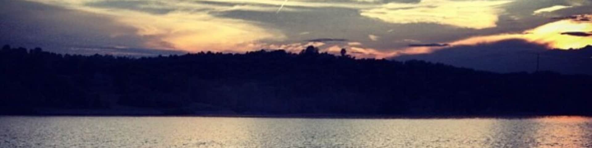 Um lago calmo sob um céu ao pôr do sol, a silhueta de uma pessoa e uma linha de árvores ao longe.