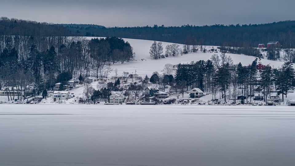 A snow-covered landscape with a cluster of houses near a frozen lake, surrounded by trees and hills.