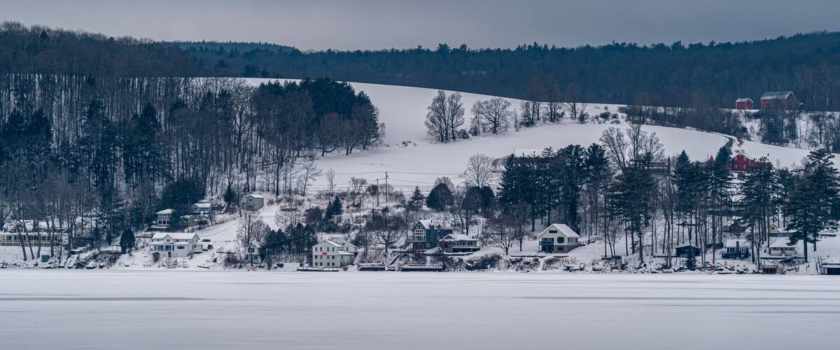 A snow-covered landscape with a cluster of houses near a frozen lake, surrounded by trees and hills.