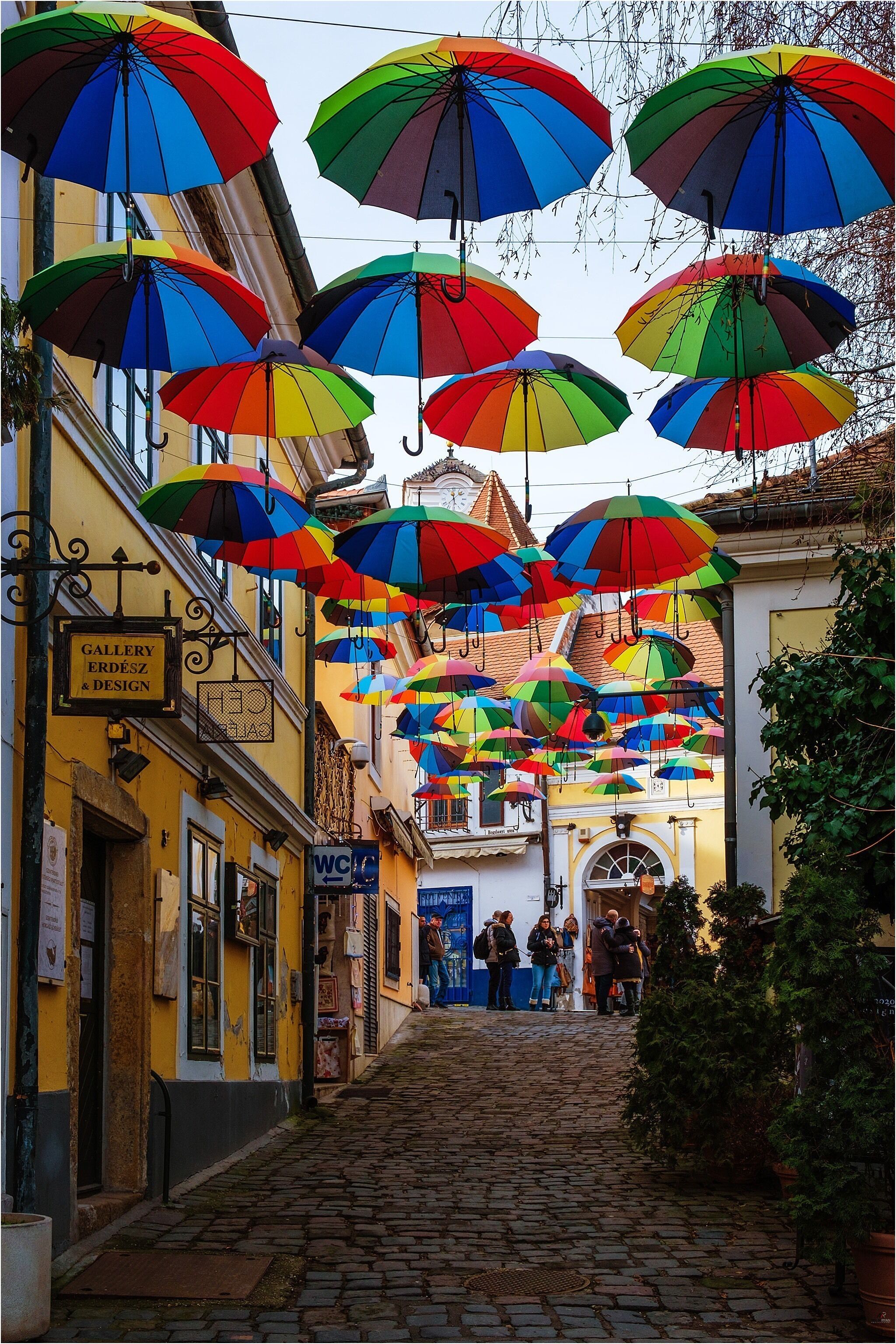 Una calle empedrada con coloridos paraguas colgando sobre ella y un letrero para una galería y tienda de diseño.