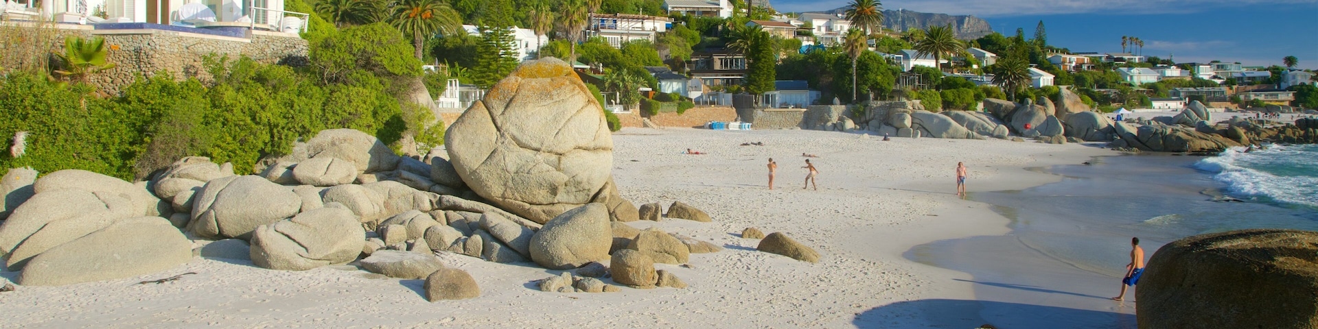 A beach with large rocks, people sunbathing, and houses in the background.