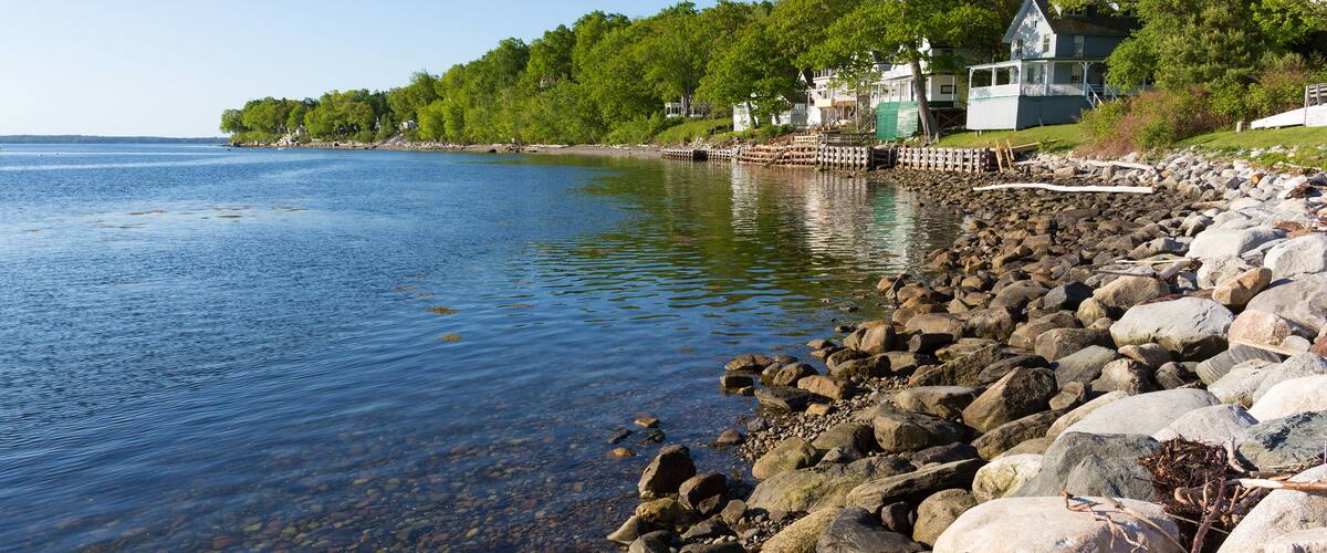 A rocky shoreline with clear water, a wooden dock, and houses with white trim and green accents.
