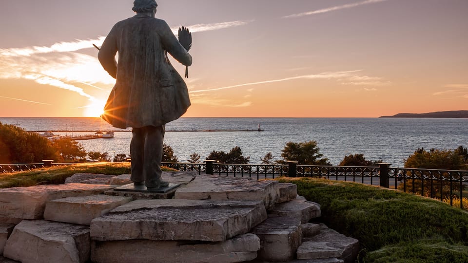 A statue overlooking a sunset over a body of water with a pier in the distance.