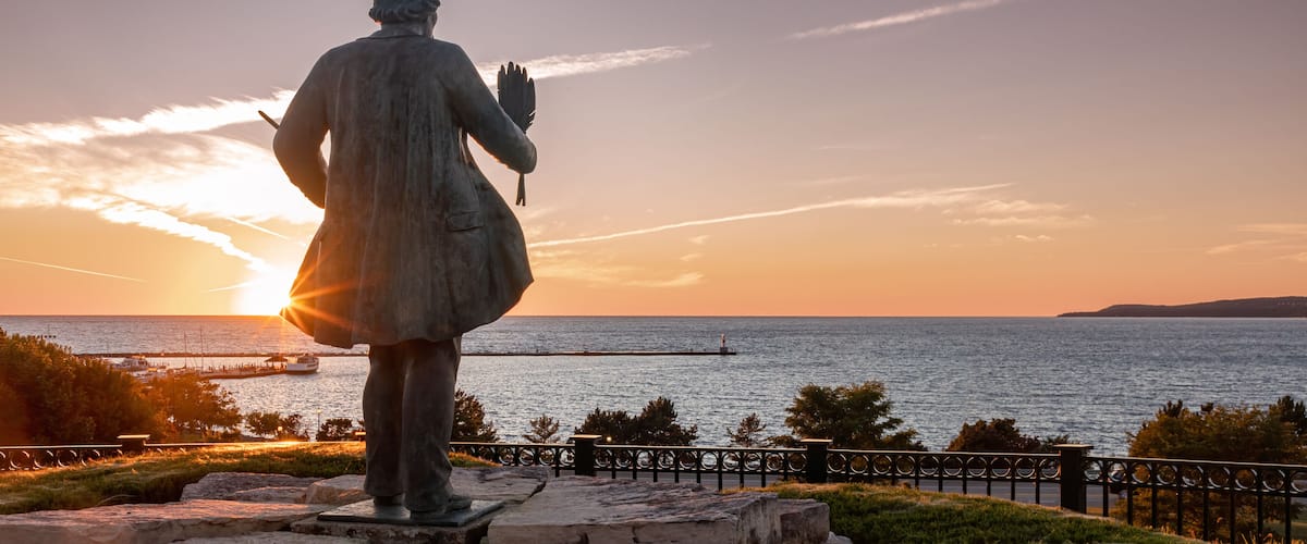 A statue overlooking a sunset over a body of water with a pier in the distance.