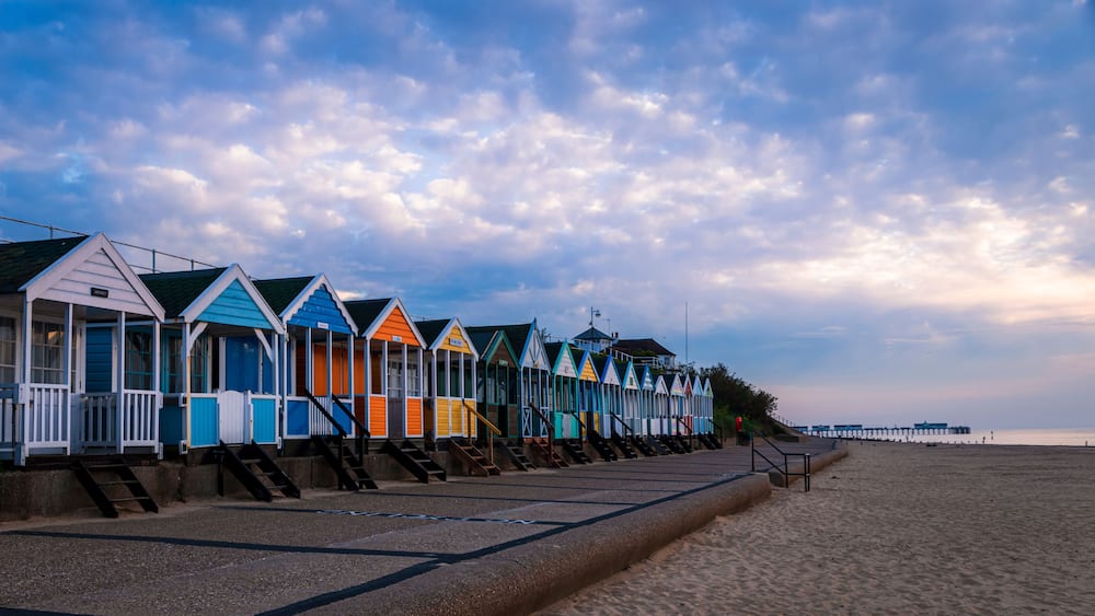 Une rangée de cabanes de plage colorées s’étend le long d’une plage de sable, sous un ciel nuageux.