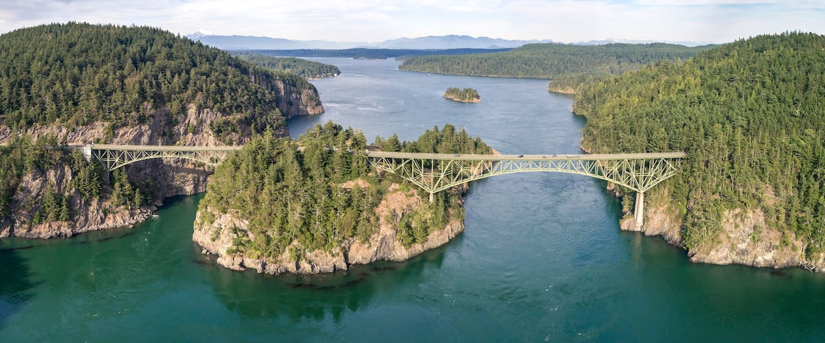 Un pont enjambant une rivière, avec des rives bordées d’arbres et des montagnes en arrière-plan.