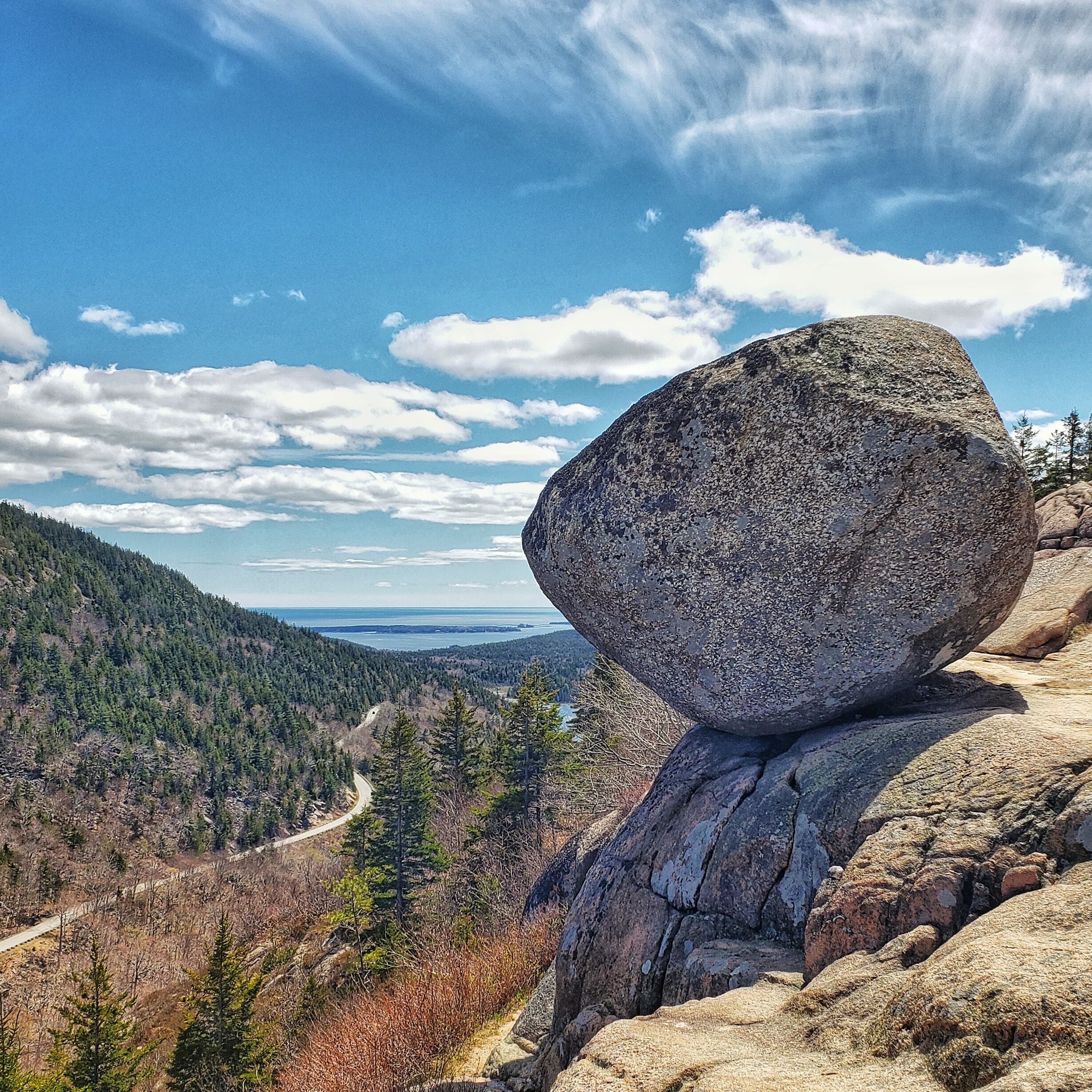 A large boulder balanced on a smaller rock, surrounded by a forested mountain landscape.