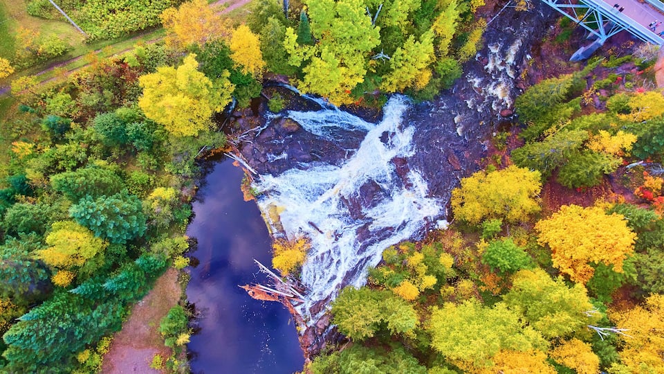 An aerial view of a river with a waterfall, surrounded by autumn-colored trees and a bridge.