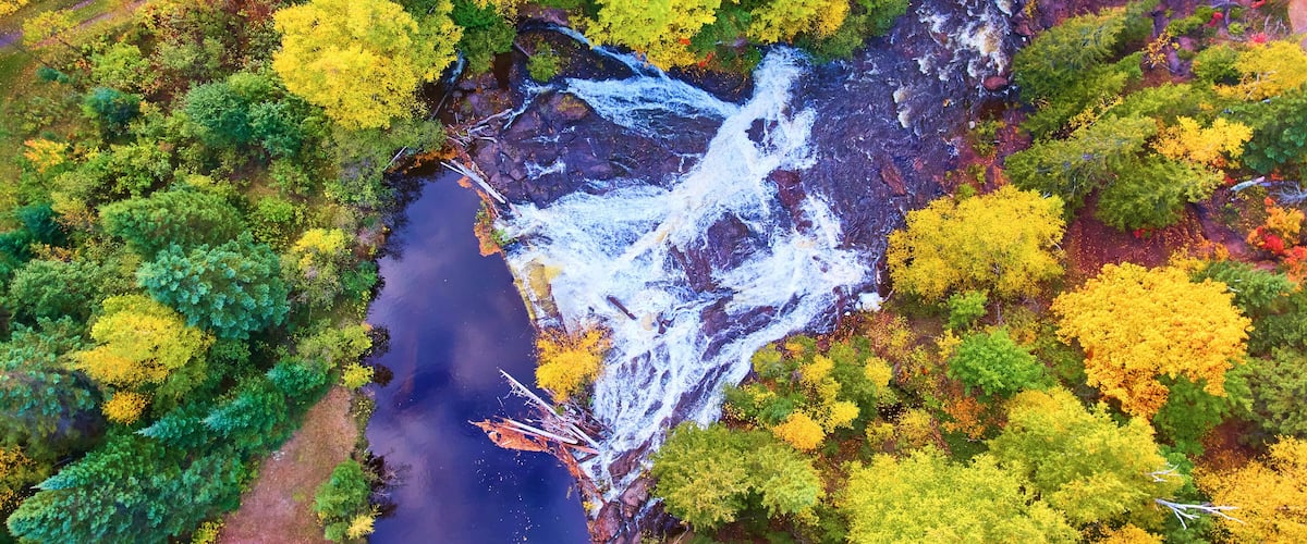 An aerial view of a river with a waterfall, surrounded by autumn-colored trees and a bridge.