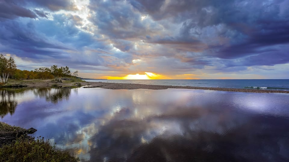 A sunset over a calm lake with a rocky shoreline and trees in the foreground.