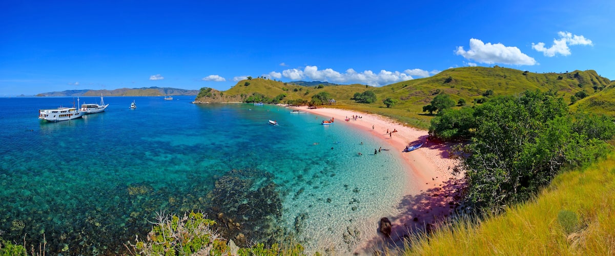 A beach with clear turquoise water, boats, and lush green hills.