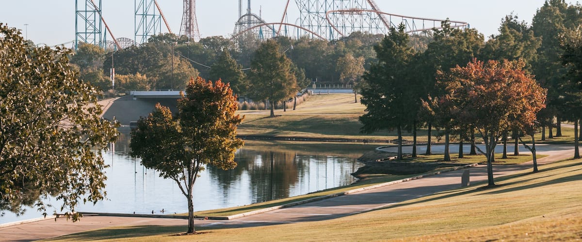 A park with a lake, trees, and roller coasters in the background.