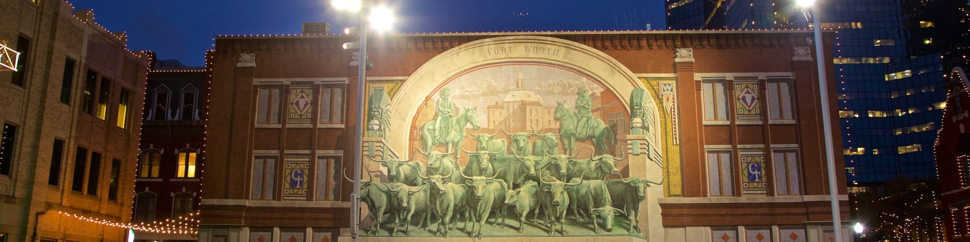 A historic building with a mural depicting a historical scene, surrounded by a plaza with fountains and modern buildings in the background.
