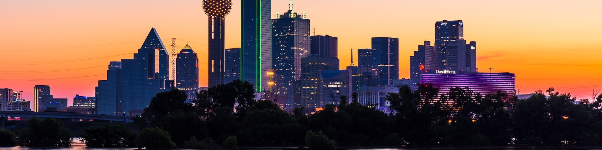 Silhueta urbana ao entardecer, com edifícios iluminados e um rio calmo em primeiro plano.