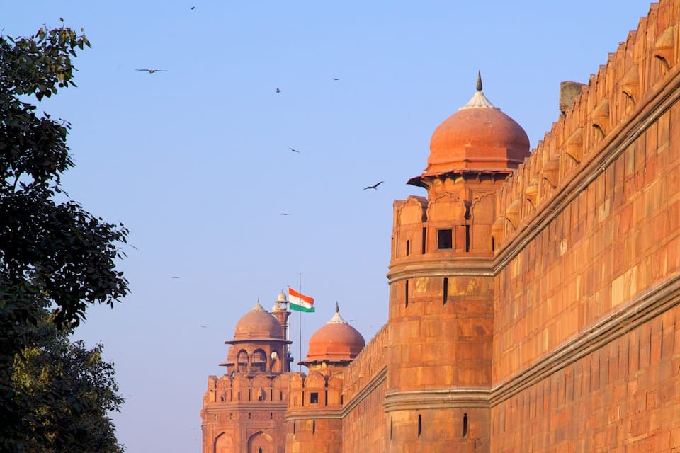 Um edifício grande e histórico com várias cúpulas e minaretes, exibindo a bandeira indiana.