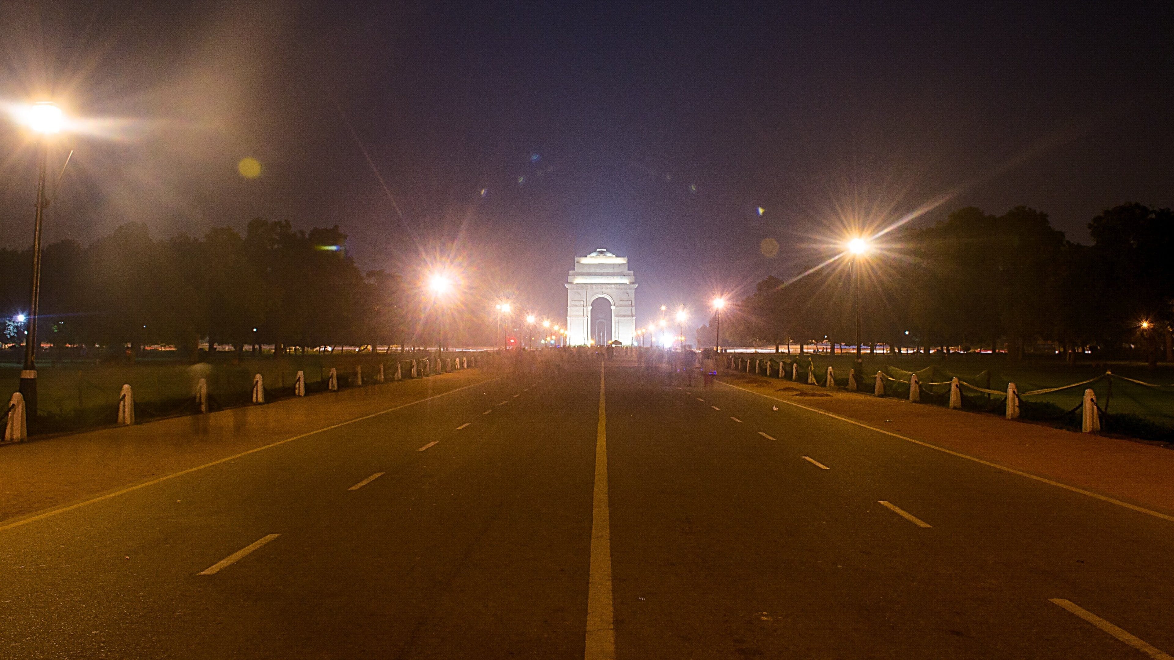 A nighttime view of a wide road leading to a prominent white archway, flanked by streetlights and barriers.