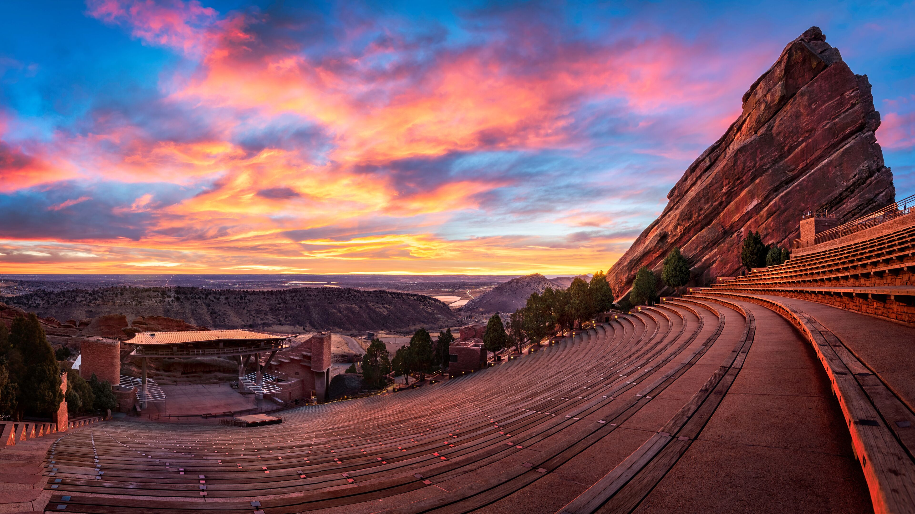 An outdoor amphitheater with tiered seating, a stage, and a large rock formation to the right.