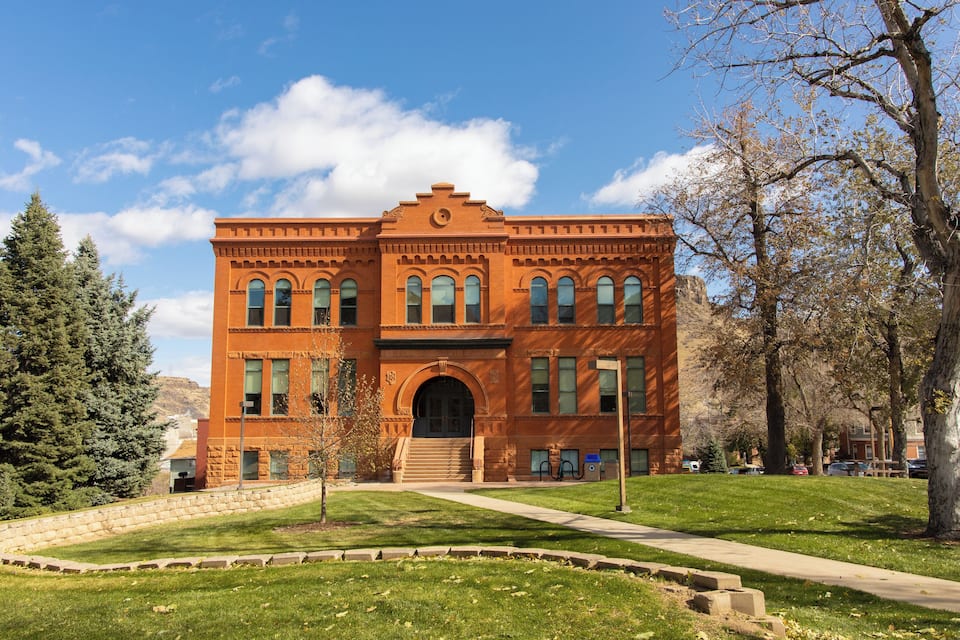 A large brick building with arched windows and a central entrance, surrounded by trees and a grassy area.