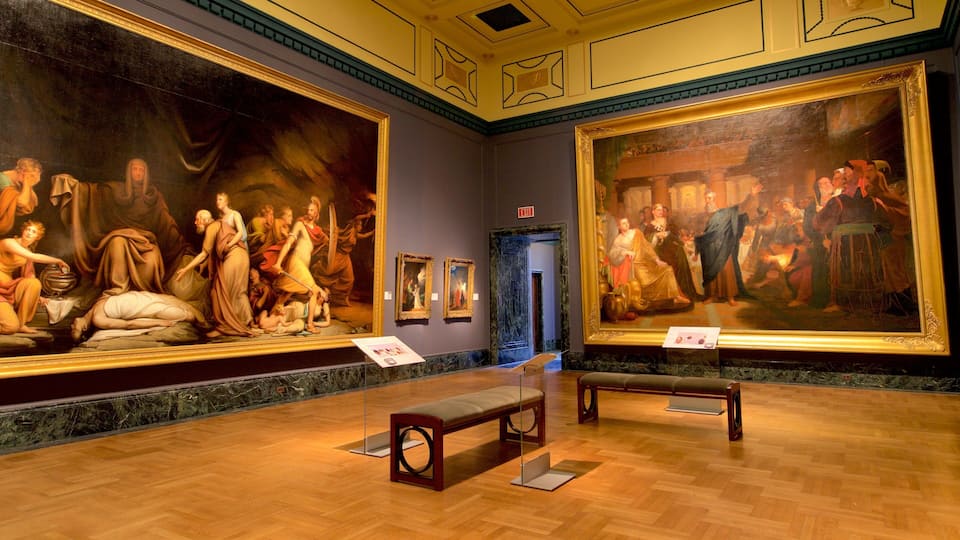 A museum interior with classical paintings on the walls and wooden benches.