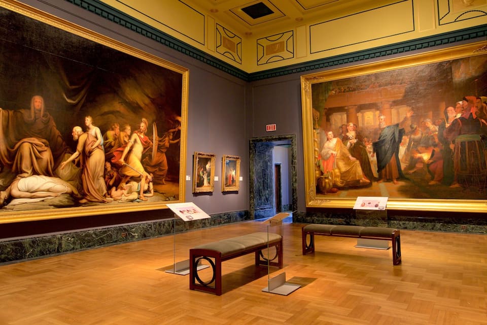 A museum interior with classical paintings on the walls and wooden benches.