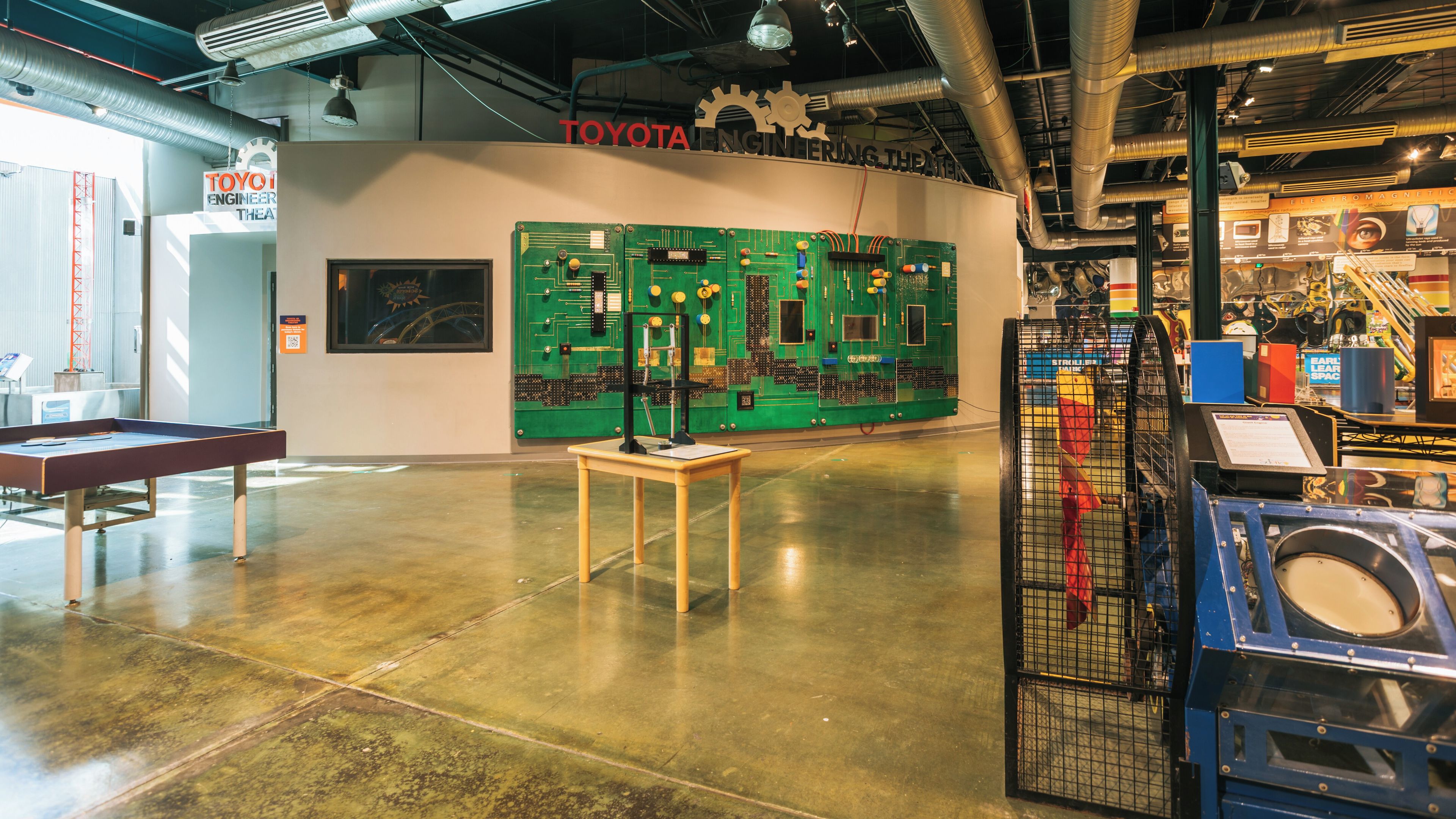 Uno spazio espositivo interno con un pannello di controllo verde, un tavolo da ping pong e diverse esposizioni meccaniche ed elettroniche.