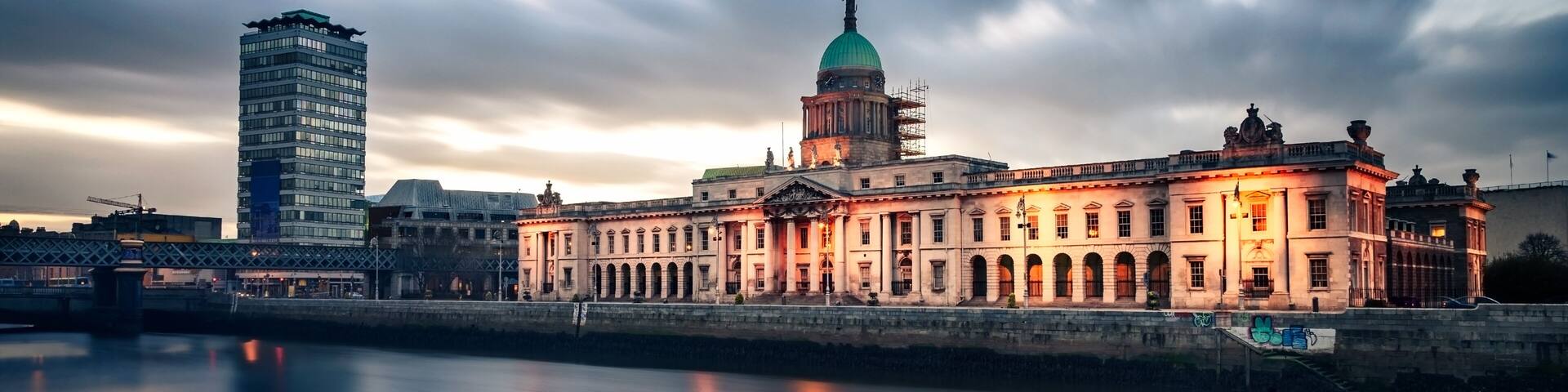 A historic building with a green dome and classical architecture, a modern skyscraper, and a calm river in the foreground.