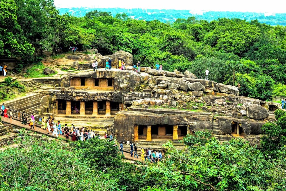 A large ancient rock structure with multiple doorways and a staircase surrounded by lush greenery.