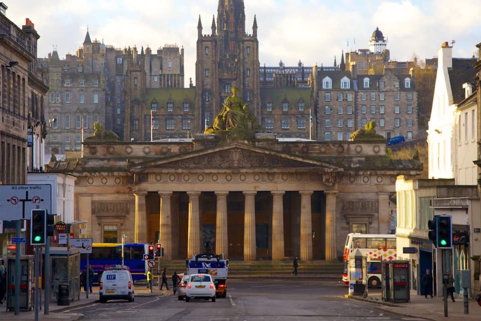 A historic building with classical architecture, including columns and statues, in an urban setting with buses and cars.